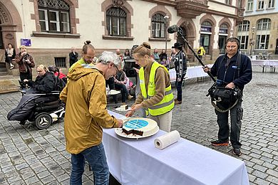 Mitarbeitenden verteilen den Kuchen an ALLE beim Protesttag zur Gleichstellung der Menschen mit Behinderungen. Dahinter ein Kamerateam des MDR