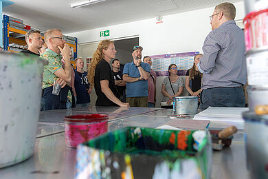 Mehrere Personen stehen in einem Praxisraum. Auf dem Tisch im Vordergrund befinden sich Farbbecher und weiteres Malmaterial. Alle Personen schauen auf einen Mann, der ihnen etwas erzählt. Dieser trägt eine Brille und ein fein gemustertes Hemd.