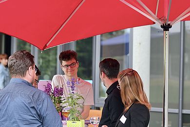 Unter einem roten Sonnenschirm kommen Teilnehmende mit den Vertreter*innen unterschiedlicher Unternehmen in Kontakt.