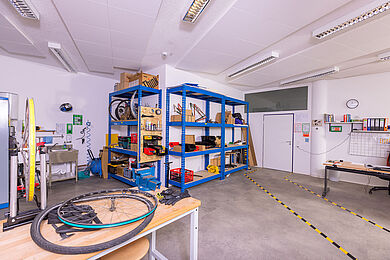 Raum der Fahrradtechnik, an der Wand blaue Metallregale mit Material und Werkzeug. Im Vordergrund ein Schreibtisch, auf dem ein Fahrradreifen und Arbeitshandschuhe liegen