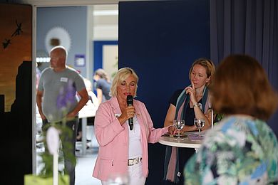 Eine Frau im rosa Blazer meldet sich zu Wort. Sie steht an einem weißen Stehtisch und spricht ins Mikrofon. Die Umherstehenden hören ihr zu.