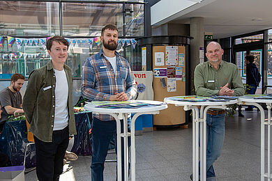 Drei Männer an Stehtischen. Alle schauen freundlich in die Kamera und haben Infomaterial vor sich ausgebreitet.
