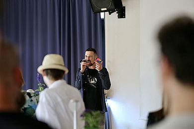 Sayes gestikuliert beim Performen mit der linken Hand. Mit der anderen hält er das Mikrofon. Über ihm hängt ein schwarzer Lautsprecher an der Wand.