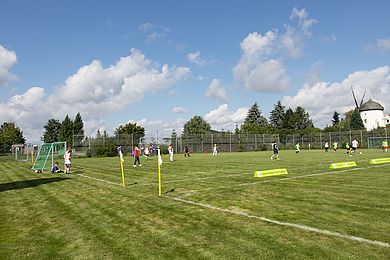 Auf dem Rasen oberhalb des Sportplatzes wird Fußball gespielt. Im Hintergrund steht die weiße Windmühle von Knauthain.