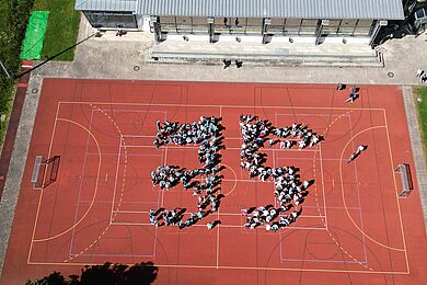 Luftaufnahme eines roten Sportplatzes vor einem Schul- oder Gebäudekomplex. Zahlreiche Personen stehen dicht beieinander und bilden gemeinsam die große Zahl „35“ in der Mitte des Platzes.