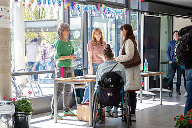 Beratungssituation im Foyer des BBW. Eine Frau im Rollstuhl und eine stehende Frau sprechen mit 2 Personen hinter einem Tisch mit Infomaterial