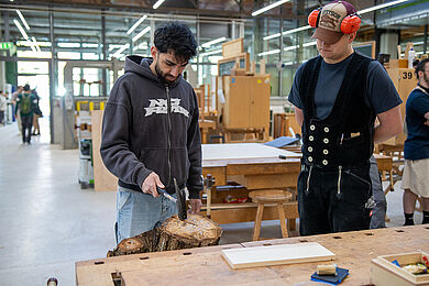 Ein Mann in Zimmermannskluft (rechts) trägt Kopfhörer auf seiner Kappe. er schaut konzentriert zu, wie ein anderer Mann mit dunklen Haaren und Bart einen Nagel mit einem Zimmermannshammer in einen Baumstamm schlägt