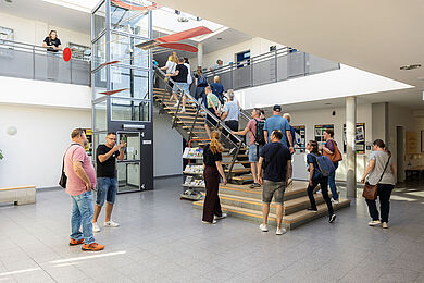 In einem Lichthof, mit einem gläsernen Fahrstuhl, gehen mehrere Personen eine Treppe nach oben.