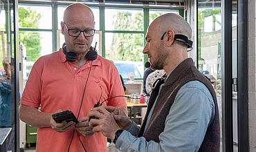 Ein Mann (rechts) erklärt einem weiteren Mann in korallfarbenen Tshirt die neue Technologier
