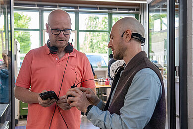 Ein Mann (rechts) erklärt einem weiteren Mann in korallfarbenen Tshirt die neue Technologier