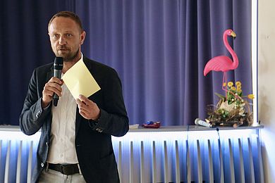 Tobias Schmidt, Hauptgeschäftsführer der BBW-Leipzig-Gruppe, begrüßt alle Teilnehmenden beim Netzwerkdinner im Freizeithaus. Er trägt ein weißes Hemd und eine helle Jeans mit schwarzem Gürtel. Auf dem Fensterbrett im Hintergrund steht ein pinker Flamingo als Dekoration.
