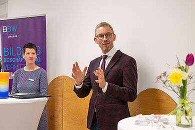 Mittig Prof. Dr. med. Fuchs bei einer Rede. Herr Fuchs trägt einen dunklen Anzug und gestikuliert mit beiden Händen. Links von ihm steht Lea Beuter an einem Stehtisch und hört ihm zu. Rechts auf einem Stehtisch ein unscharf abgebildeter Blumenstrauß.