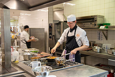 Im Vordergrund kocht ein Azubi, im Hintergrund läuft ein anderer geschäftig vorbei
