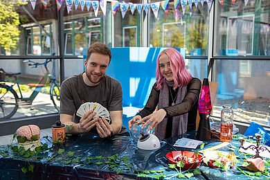 Ein Mann (links) und eine Frau an einem Tisch. Der Mann lächelt und hält Karten aufgefächert in den Händen. Die Frau hat rosa Haare und lächelt ebenfalls in die Kamera. Sie hält eine Glaskugel.