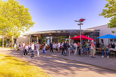 Viele Personen stehen vor einem Gebäude und unterhalten sich ausgiebig. Außerdem sind zwei Sonnenschirme in rot und blau aufgestellt, die Sonne scheint.