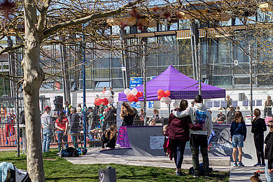 Blick auf die Sporthalle und den Sportplatz, auf dem sich viele Menschen tummeln. An einem lila Pavillon sind Luftballons in rot und weiß angebracht. 