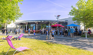 Viele Personen stehen vor einem Gebäude und unterhalten sich ausgiebig. Außerdem ist ein Sonnenschirm in rot aufgestellt, die Sonne scheint. Das Gebäude grenzt an eine schöne grüne Wiese, auf welcher im Vordergrund ein pinkfarbener Liegestuhl steht.
