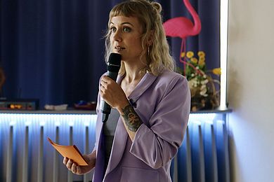 Carina Krause führte als Moderatorin durch die Veranstaltung. Sie trägt einen fliederfarbenen Blazer, ein Tattoo am linken Unterarm und zwei Lippenpiercings. Ihr blondes Haar ist wellig und reicht bis zu den Schultern.