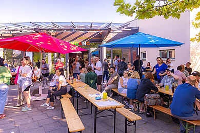 An mehreren Biertischgarnituren, die vor einem Gebäude stehen, sitzen Personen. Sie unterhalten sich freudig und trinken verschiedene Getränke. Zum Schutz gegen die Sonne sind zwei Sonnenschirme in rot und blau aufgestellt. Ringherum stehen und unterhalten sich weitere Personen.