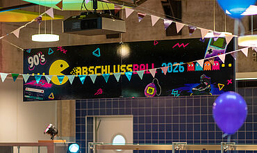 Großes Banner im Stile der 90er mit der Aufschrift "Abschlussball 2026", Davor bunte Girlanden und Luftballons