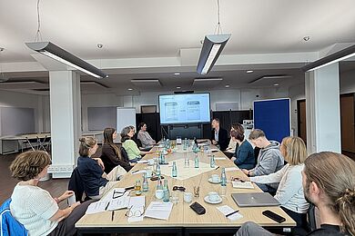 11 Personen sitzen um einen länglichen Tisch herum in einem großen Büroraum. Sie schauen auf eine digitale Tafel auf der eine Tabelle aufgezeigt wird.