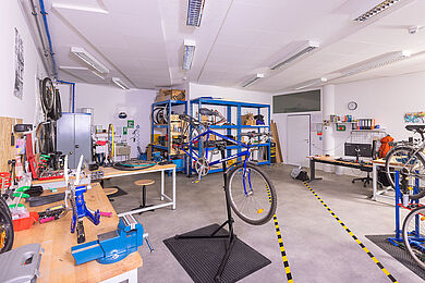 Räumlichkeiten der Fahrradwerkstatt. links im Bild eine Werkbank mit verschiedenen werkzeugen, in der Mitte ein blaues Fahrrad ohne Hinterrad auf einem Montageständer. Im Hintergrund Regale mit Material und Werkzeug sowie Arbeitsbereiche und Schreibtische