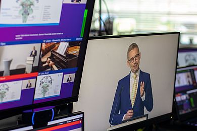 Auf einem Bildschirm sieht man Prof. Dr. Michael Fuchs vom Universitätsklinikum Leipzig. Über seinem weißen Hemd trägt er eine gelbe Krawatte und ein blaues Sakko. Markant ist seine Brille, die einen schwarzen Rahmen hat.