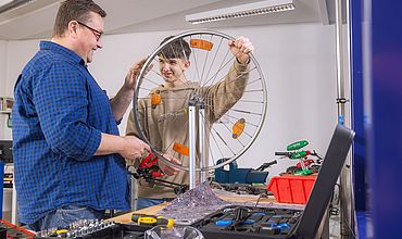 Diakonie am Thonberg Teilnehmer*in der BvB-P-Maßnahme bei der Arbeit. Ein*e Mitarbeiter*in in blauem Hemd begleitet einen Teilnehmenden mit dunklen kurzen Haaren und braunem Sweatshirt bei der Reparatur eines Fahrrads.