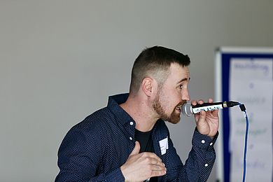 Der MC, politische Aktivist, Freestyler und Lyriker Gregor Zocher alias Sayes performte live beim Netzwerkdinner. Er trägt einen Bart, ein blaues Hemd mit kleinen weißen Punkten und darunter ein schwarzes T-Shirt mit Aufdruck. Seine braunen Haare sind an den Seiten kurz rasiert.