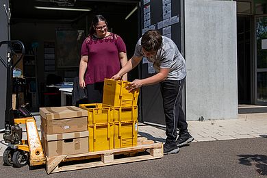 Zwei Auszubildende beladen eine Palette mit Kisten.