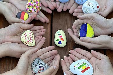 Zum Thema „Respekt und Toleranz“ haben die Teilnehmenden Steine bemalt. Auf dem Bild sieht man kreisförmig angeordnete Hände, die ihre kreativen Kunstwerke in die Kamera halten. Gern gewählte Motive sind Länderflaggen und Herzchen.