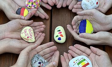 Zum Thema „Respekt und Toleranz“ haben die Teilnehmenden Steine bemalt. Auf dem Bild sieht man kreisförmig angeordnete Hände, die ihre kreativen Kunstwerke in die Kamera halten. Gern gewählte Motive sind Länderflaggen und Herzchen.