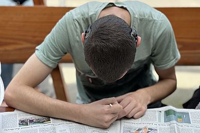 Ein Teilnehmender steht über den mit Zeitungen bedeckten Tisch gebeugt und mal mit einem Pinsel. Hinter seinen Ohren sind zwei Hörgeräte zu sehen.