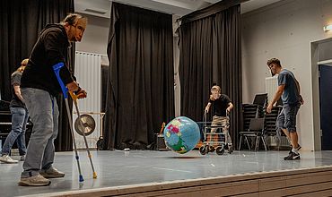 Performance auf der Bühne bei der Theatershow - vier Personen spielen Theater. Sie haben Masken auf. Eine Person vorne links läuft mit Krücken. In der Mitte dotzt ein Wasserball bedruckt mit der Erdkügel auf.