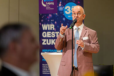 Im Fokus des Bildes steht der Moderator des Abends, Udo Färber. Er trägt einen Anzug in Altrosa und eine blau-weiß gestreifte Krawatte. Er trägt eine Brille und steht vor einem Banner, das auf das 35. Jubiläum des BBW hinweist.