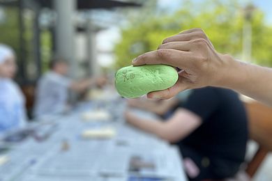 Im Hintergrund sieht man verschwommen mehrere Leute an einem Tisch sitzen, auf dem einige Zeitungen zum Schutz vor Farbflecken ausgebreitet wurden. Im Vordergrund hält jemand einen hellgrün bemalten Stein in die Kamera, auf den bereits ein Stichmännlein zu sehen ist.