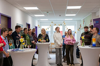 Blick in den Raum auf die Gäste der Eröffnungsfeier, die sich im Halbkreis aufgestellt haben. Alle blicken freudig auf Dr. Ronny Valdorf, rechts im Bild, der gerade das Wort ergriffen hat.