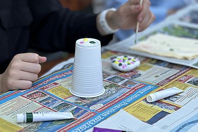 Auf einer Zeitung steht ein umgestülpter Plastikbecher, der als Farbpalette dient. Außerdem sind die Hände der malenden Person zu sehen, die mit einem Pinsel grüne, gelbe und pinke Punkte auf einen Stein aufgetragen hat.