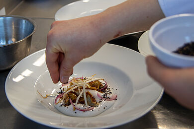 Ein Teller wird angerichtet, eine Hand streut gerade ein schwarzes Gewürz auf die Speisen