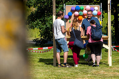 Auf einer Wiese steht ein Stand, an dem eine Wand aus bunten Luftballons befestigt ist. Vor dem Stand stehen drei Personen, die der Person zugewandt sind, die hinter dem Stand steht. Diese scheint ihnen etwas zu erklären.