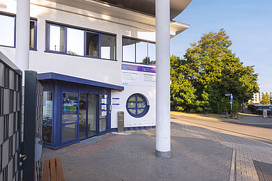 Blick auf den haupteingang der Witzgallstraße 6 von links. Der Eingang ist blau eingefasst und mit einer Glasschiebetür. Daneben ist ein rundes Fenster. Davor eine Säule, die das Vordach trägt. 